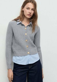 Grauer Cardigan mit geripptem Design, verziert mit goldenen Knöpfen, über einem hellblauen gestreiften Hemd getragen. Stilvolles, lässiges Design mit figurbetonter Schnittführung.