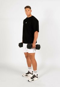 T-shirt noire surdimensionnée, shorts blancs, tenant des haltères noirs. Baskets blanches texturées avec des accents rouges et noirs. Design minimaliste.