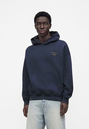 Ung mand iført en marineblå hættetrøje med en frontlomme og lyseblå jeans, stående foran en ensfarvet hvid baggrund.