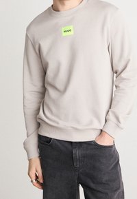 Lichtgrijze sweater met een ronde hals en geribde manchetten, met een vierkante neon groene logopatch met de tekst "HUGO" op de borst.