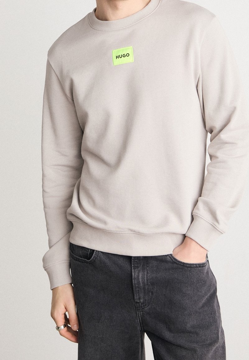 Lichtgrijze sweater met een ronde hals en geribde manchetten, met een vierkante neon groene logopatch met de tekst "HUGO" op de borst.