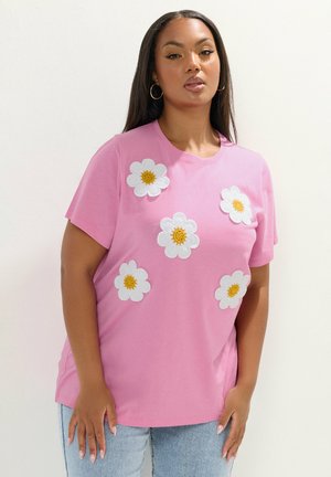 Kvinde med langt mørkt hår iført guldringeøreringe og en lyserød t-shirt dekoreret med fem hvide og gule margueritblomster.