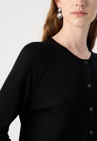 Svart cardigan med rund hals, strukturert strikket stoff og knappelukking foran. Har raglanermer og svarte knapper.