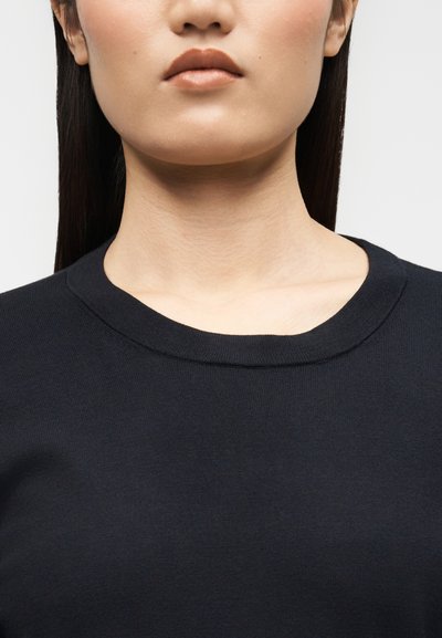 Gros plan sur le bas du visage et le cou d'une personne portant un t-shirt noir à col rond, avec des cheveux raides et foncés visibles sur les côtés.