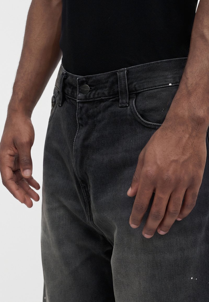 Close-up of a person wearing faded black jeans with a button closure, showing hands resting beside the body against a plain background.
