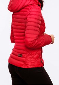 Eine Frau trägt eine enganliegende rote gesteppte Jacke mit Kapuze, steht seitlich und hält ihre rechte Hand in der Nähe des Reißverschlusses der Jacke.