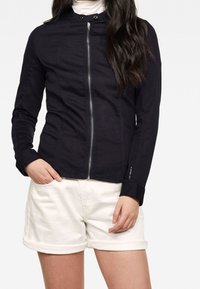 Giacca nera aderente con chiusura a zip, collo alto e maniche lunghe. Indossata sopra un dolcevita bianco e abbinata a pantaloncini bianchi.