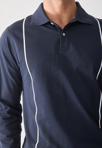 Mornarska polo majica dugih rukava s klasičnim kragnom, bijelim bočnim prugama i teksturiranim materijalom. Placket s tri gumba na vratu.