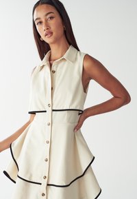 Donna con lunghi capelli scuri che indossa un vestito beige senza maniche con rifiniture e bottoni neri, in posa con una mano sui fianchi su uno sfondo bianco.