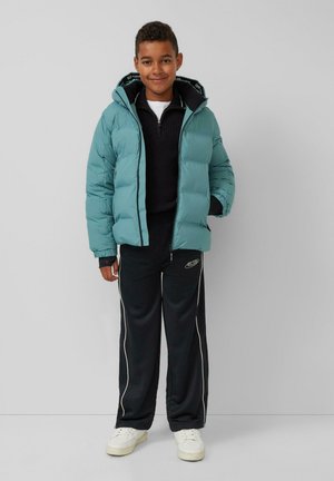 Giacca a vento teal con cappuccio, chiusura con zip e tasche laterali, indossata sopra un maglione nero a maglia e pantaloni della tuta neri con strisce bianche e logo.