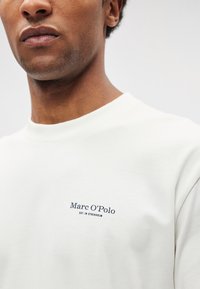 Gaiši krēmīga kokvilnas džemperis ar apaļu kakla izgriezumu, uz kreisās krūšu daļas tumši zili izšūts teksts "Marc O'Polo EST. IN STOCKHOLM".
