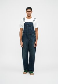 Dunkelblaue Jeans-Latzhose mit weitem Bein, Vordertasche und verstellbaren gestreiften Trägern. Trägt ein weißes T-Shirt und grüne Turnschuhe.