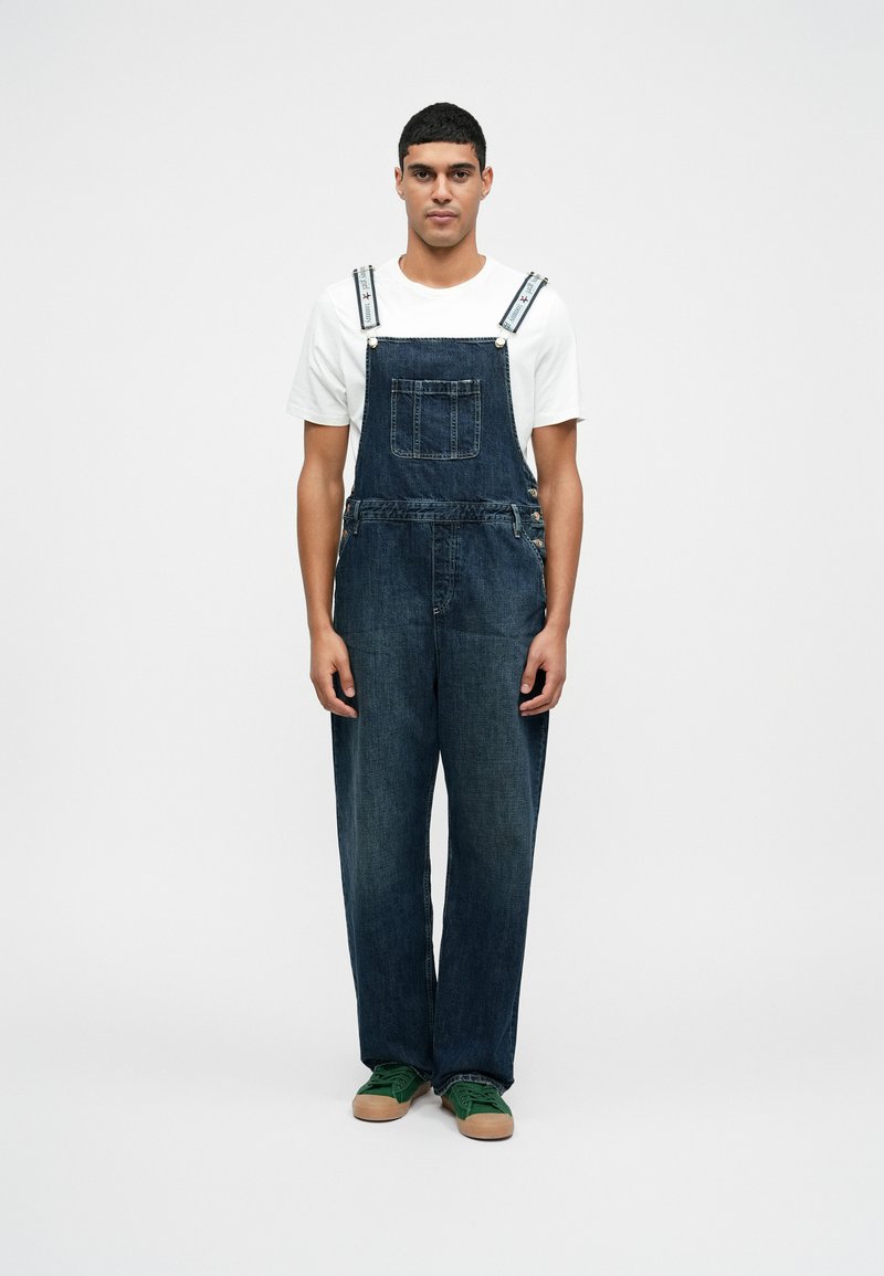 Dunkelblaue Jeans-Latzhose mit weitem Bein, Vordertasche und verstellbaren gestreiften Trägern. Trägt ein weißes T-Shirt und grüne Turnschuhe.