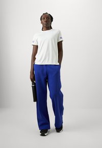 Valge lühikeste varrukatega T-särk külgsuunaliste lõigetega ja sinised laiad püksid; seisab musta veepudeli kõrval minimalistlikus keskkonnas.