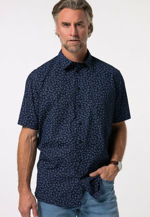 Man draagt een marineblauw overhemd met korte mouwen en een klein bloemenpatroon en lichtblauwe spijkerbroek, hand in zak.