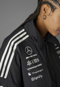 Crni windbreaker s bijelim prugama. Ima logotipe: Mercedes, AMG, UBS, Crowdstrike, Snapdragon. Uključuje visoki ovratnik i patentne zatvarače.