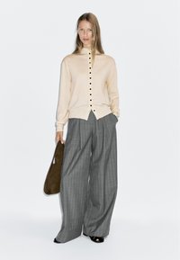 Cardigan beige avec des boutons noirs, associé à un pantalon large à rayures gris. Accessoire : sac à épaule vert foncé. Chaussures entièrement noires.