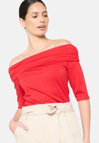 Top rosso a spalle scoperte con dettaglio arricciato, maniche a tre quarti e tessuto morbido e liscio. Abbinato a pantaloni beige con cintura.