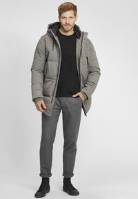 Doudoune grise avec capuche, fermeture éclair et poches latérales ; portée sur un t-shirt noir, associée à un pantalon gris à motifs et des baskets noires.