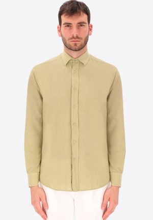 Uomo con capelli corti e scuri che indossa una camicia beige a maniche lunghe abbottonata e pantaloni bianchi, in piedi davanti a uno sfondo chiaro e semplice.