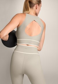 Könnyű, testhezálló edző szett tompa taupe színben. A sportmelltartón kulcslyuk kivágás található; a leggings sima textúrával rendelkezik, minimális részletezéssel.