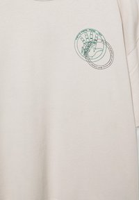 Chemise couleur crème avec un graphisme d'une main et deux motifs circulaires superposés contenant du texte sur la partie supérieure gauche de la poitrine.