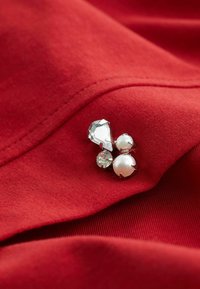 Drie kleine decoratieve edelstenen, waaronder twee parels en één heldere facette-gem, vastgenaaid op rode stof met zichtbare stiksels.