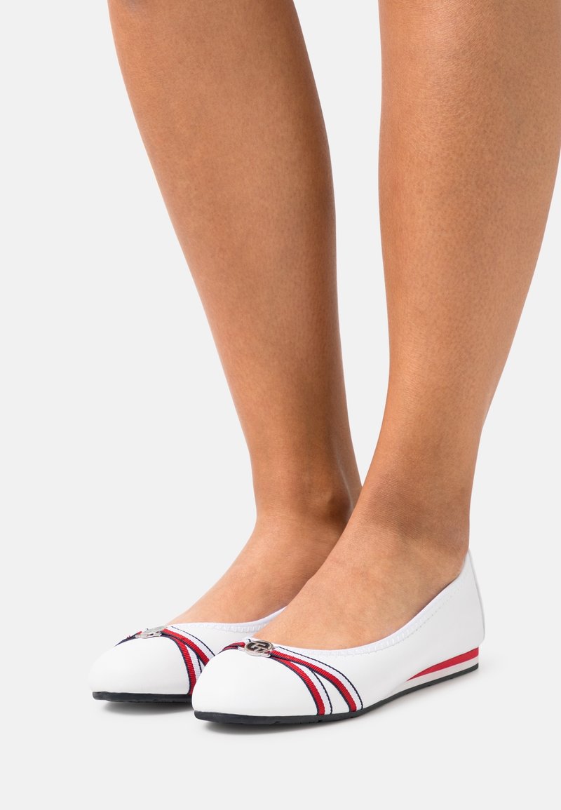 Pieds portant des ballerines blanches avec des rayures rouges et noires et un petit logo métallique sur les orteils, présentés sur un fond clair uni.