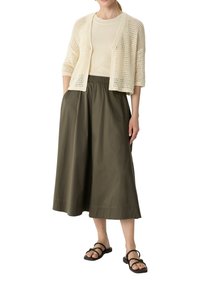 Hellbeige gestrickte Strickjacke über einem cremefarbenen Top, kombiniert mit einem olivgrünen, knielangen Rock und schwarzen Riemchensandalen.