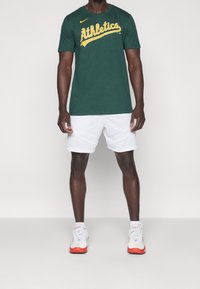 Zelené tričko Nike s nápisem „Athletics“ žlutou barvou, v kombinaci s bílými šortkami. Model má na nohou bílé a červené tenisky.