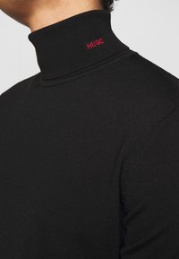 Pullover nero con collo alto in maglia a coste e design aderente. Presenta un piccolo logo rosso "HUGO" sul colletto.