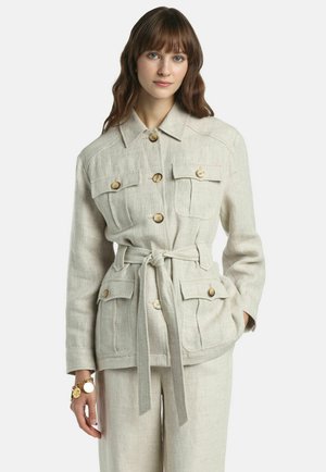 Nő bézs színű, övvel ellátott lenvászon dzsekiben, gombos zsebfedőkkel és hozzáillő bő szárú nadrágban, egyszínű háttér előtt áll.