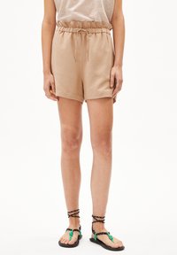 Beige shorts met een elastische tailleband en trekkoord, met een gladde textuur en zijzakken. Gecombineerd met zwart-groene sandalen met bandjes.