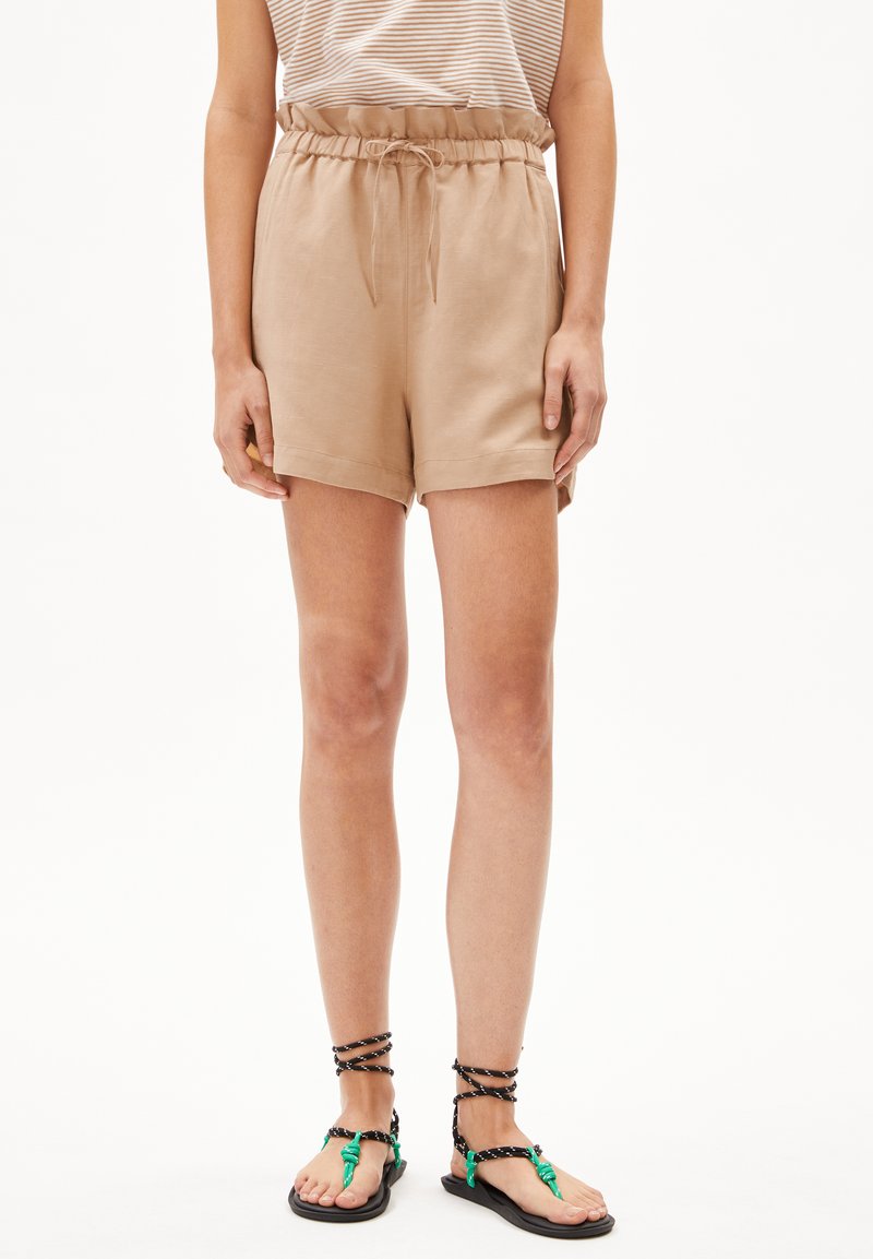 Beige shorts met een elastische tailleband en trekkoord, met een gladde textuur en zijzakken. Gecombineerd met zwart-groene sandalen met bandjes.