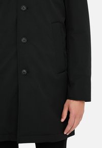 Schwarze Außenjacke mit glatter Textur, ausgestattet mit einem Knopfverschluss, Seitentaschen und geradem Saum, präsentiert mit einer Hand auf dem Stoff.