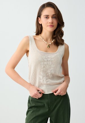 Jonge vrouw die een beige mouwloze gebreide top met bloemenpatroon en groene broek draagt, geaccentueerd met een schelpenketting, houdt de zoom van de top vast.