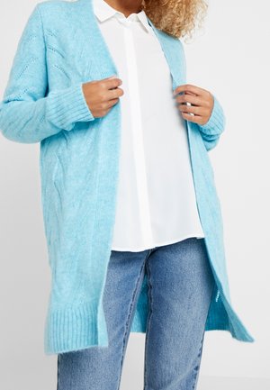 Osoba nosící světle modrý pletený kardigan přes bílou košili s knoflíčky a modré džíny, upravující přední okraje kardiganu.