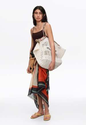 Mujer con camiseta sin mangas marrón oscuro y falda cruzada multicolor, llevando un gran bolso blanco tejido y sandalias amarillas.