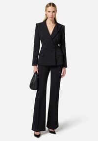 Blazer nero doppiopetto con rever a punta, abbinato a pantaloni su misura coordinati. La modella tiene una borsa nera texture.