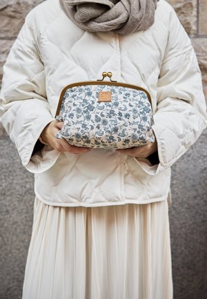 Person i hvid quiltet jakke og beige tørklæde holder en clutch med blomsterprint og guldramme mod en baggrund af stenmuret væg.