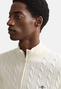 Kremfarvet kabelstrikket sweater med høj ribstrikket krave og lynlås foran. Har diskret logo-broderi på venstre bryst.