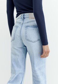 Persoană purtând blugi de denim albastru deschis și un top cu mâneci lungi bleu marin, văzută din spate, cu buzunare vizibile și etichetă de brand pe talie.