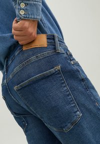 Sötétkék farmernadrág sima textúrával. Bőrből készült folttal a hátsó zseben és standard varrás részletekkel rendelkezik.
