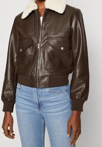 Giacca bomber in pelle marrone con colletto in shearling, polsini e orlo a costine, due tasche frontali e chiusura con zip. Si abbina a jeans azzurri chiari.