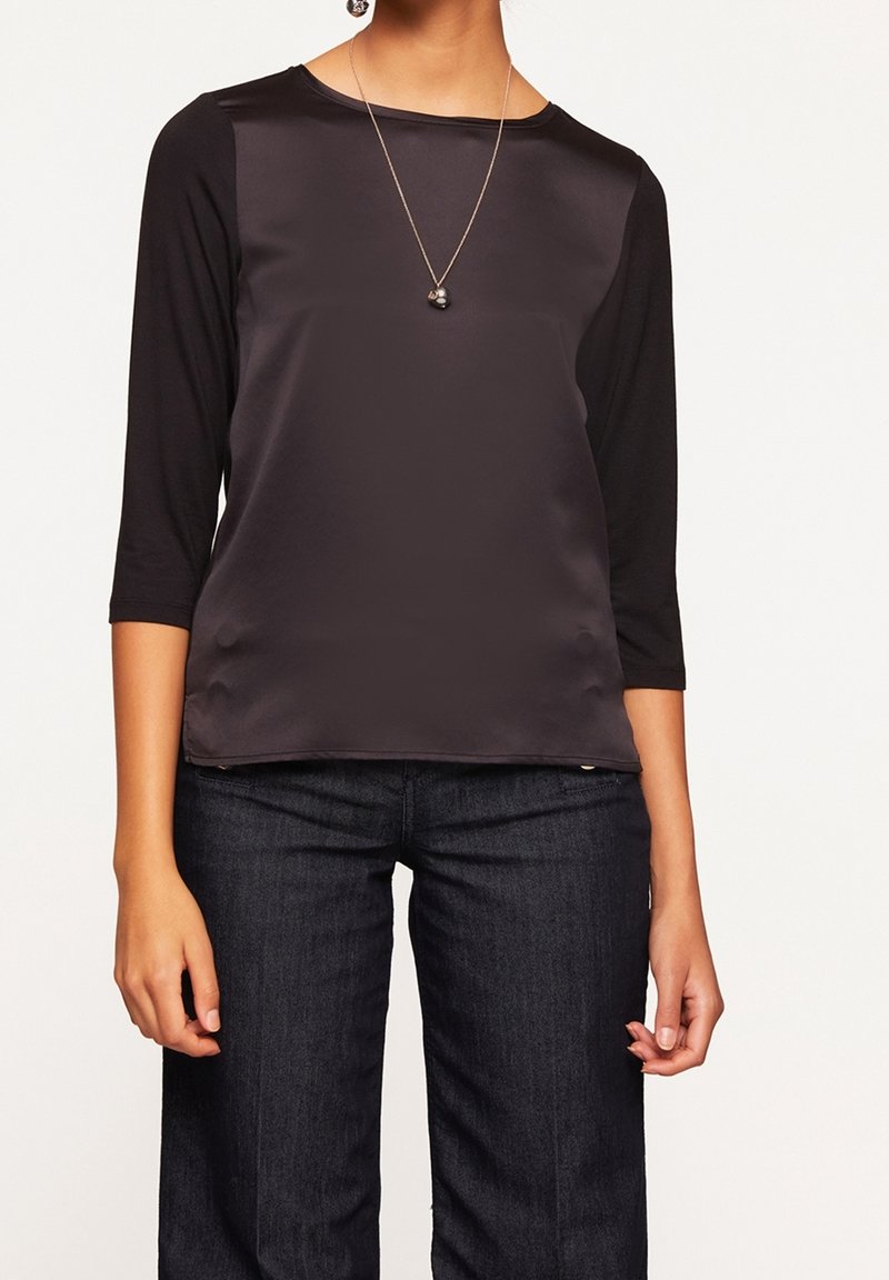 Černý dlouhý rukáv top s saténovým předním dílem a džersejovým zadním dílem. Výstřih je kulatý a jednoduchý náhrdelník doplňuje celkový vzhled. V kombinaci s tmavými džíny.