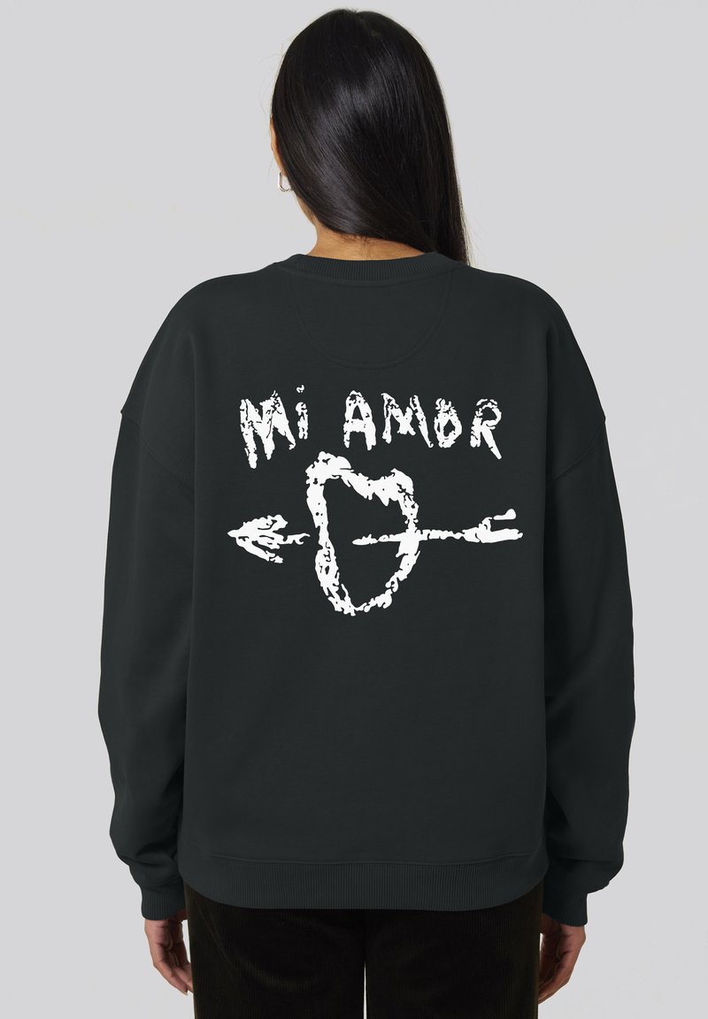 Felpa nera con una grafica bianca che presenta il testo "Mi Amor" e una freccia che attraversa un cuore sul retro. Tessuto morbido, vestibilità oversize.