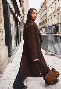 Brown wool coat with a tailored fit, wide lapels, and front pockets. Carrying a brown leopard-print handbag. Urban street setting.