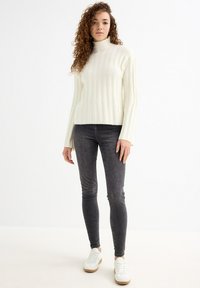 Suéter de canalé color crema con cuello alto, combinado con jeans ajustados gris oscuro y zapatillas blancas con suelas marrones. Textura suave en general.