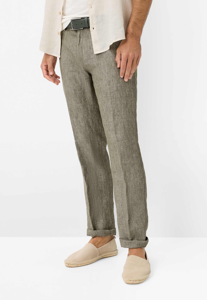 Hombre vistiendo pantalones de lino grises con puños, zapatos beige sin cordones, cinturón negro y camisa abierta beige claro sobre una camiseta blanca.