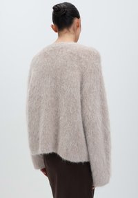 Woman with dark hair in a bun wearing a loose, fuzzy beige sweater and dark brown pants, shown from the back against a plain background.